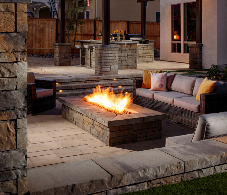 Modern patio with a rectangular stone fire pit, wicker sofas, and an outdoor kitchen in the background.