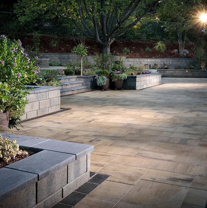 Inspiration_Patios_Image6 Spacious stone patio with raised garden beds, potted plants, and a large tree in the background.