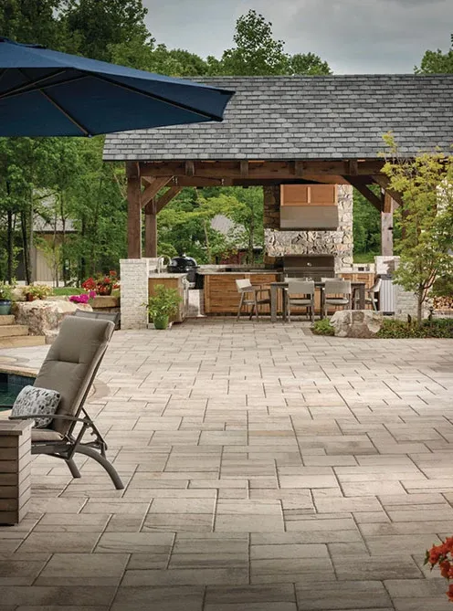 Outdoor patio with lounge chair, pavers, and covered kitchen dining area surrounded by greenery.