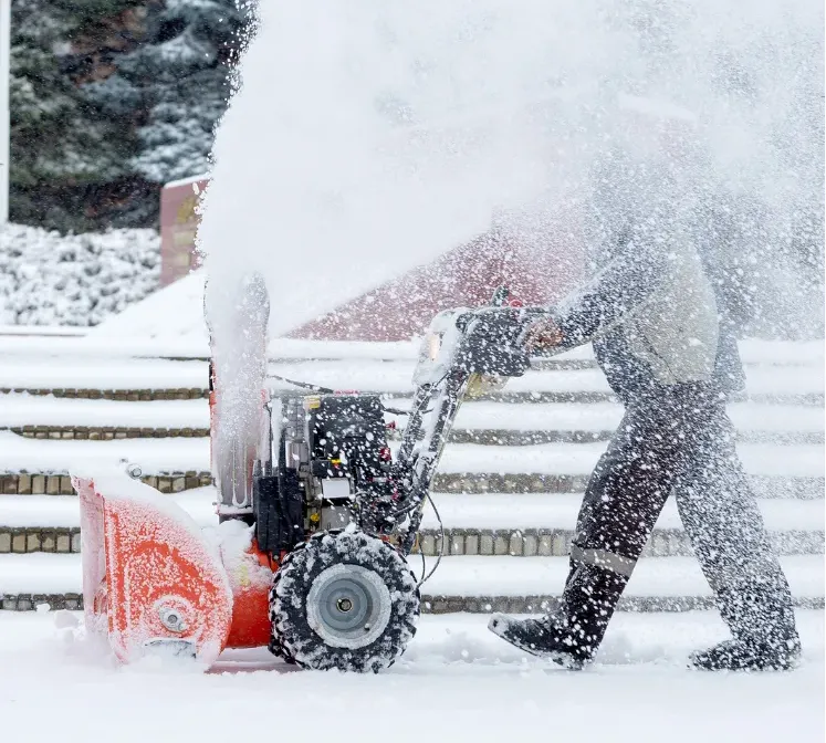 A person uses a snow blower, clearing heavy snow in front of outdoor steps on a winter day.