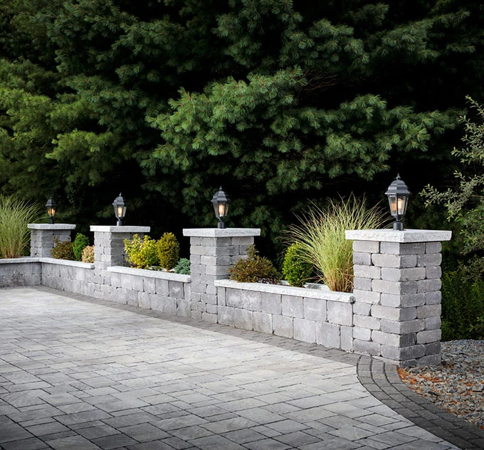 walls-bg-2 Gray stone garden wall with lanterns and plants, next to a paved driveway and tall evergreen trees.
