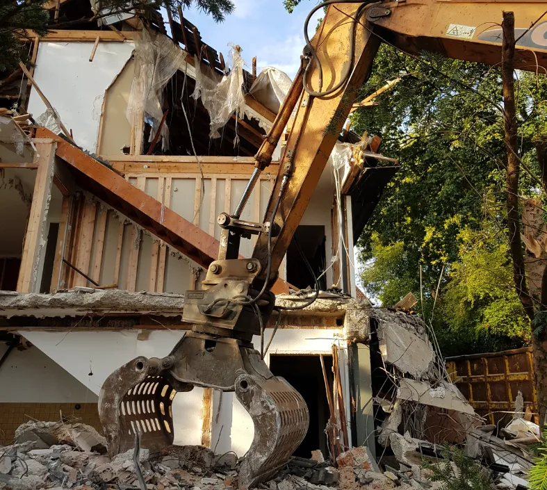 Excavator demolishing a partially collapsed house surrounded by rubble and trees.