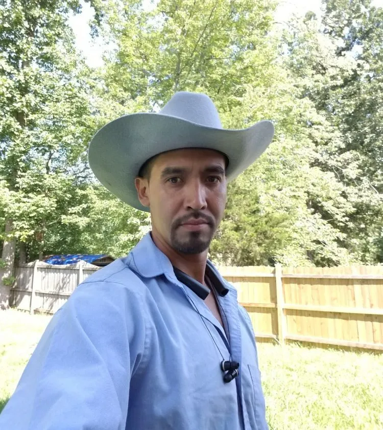 Man wearing a cowboy hat and blue shirt stands outside in a sunny backyard with trees and a wooden fence.